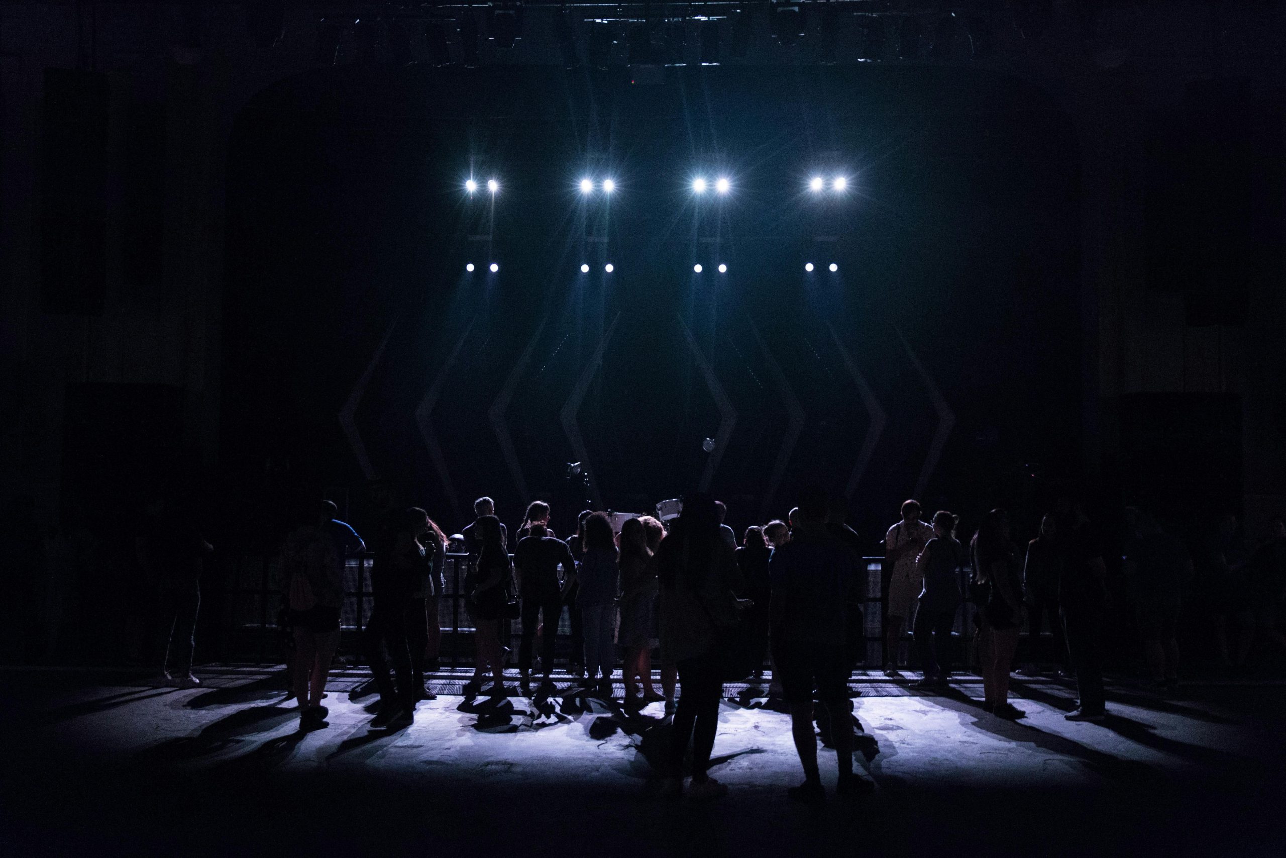 Silhouettes of a crowd gathered in front of a stage illuminated by bright lights at a live music event.